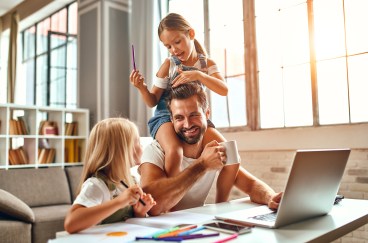Working dad with two daughters, sitting at desk with laptop, daughter on shoulders