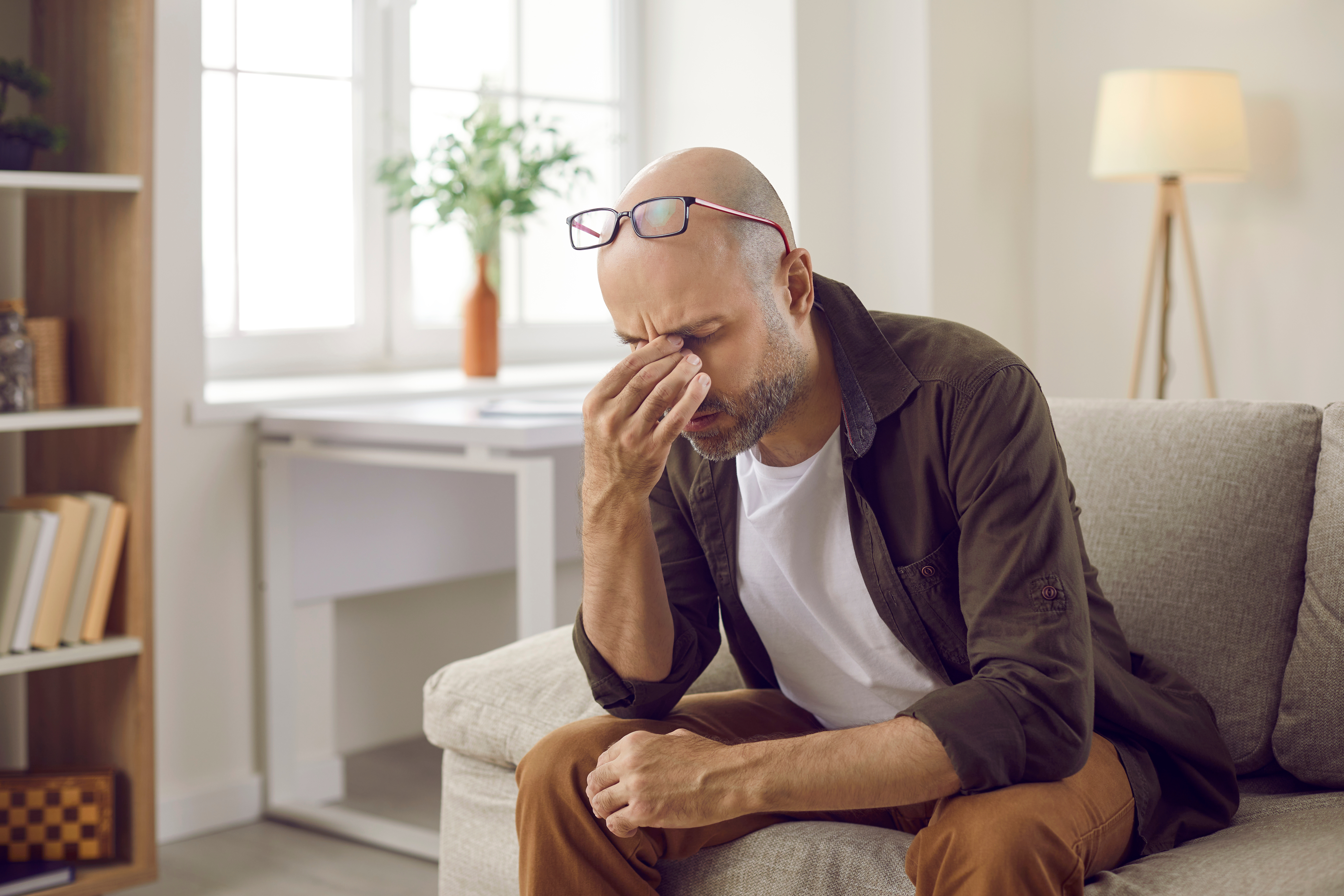 A middle-aged man pinches his nose in frustration while sitting on his couch.