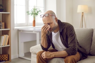 A middle-aged man pinches his nose in frustration while sitting on his couch.