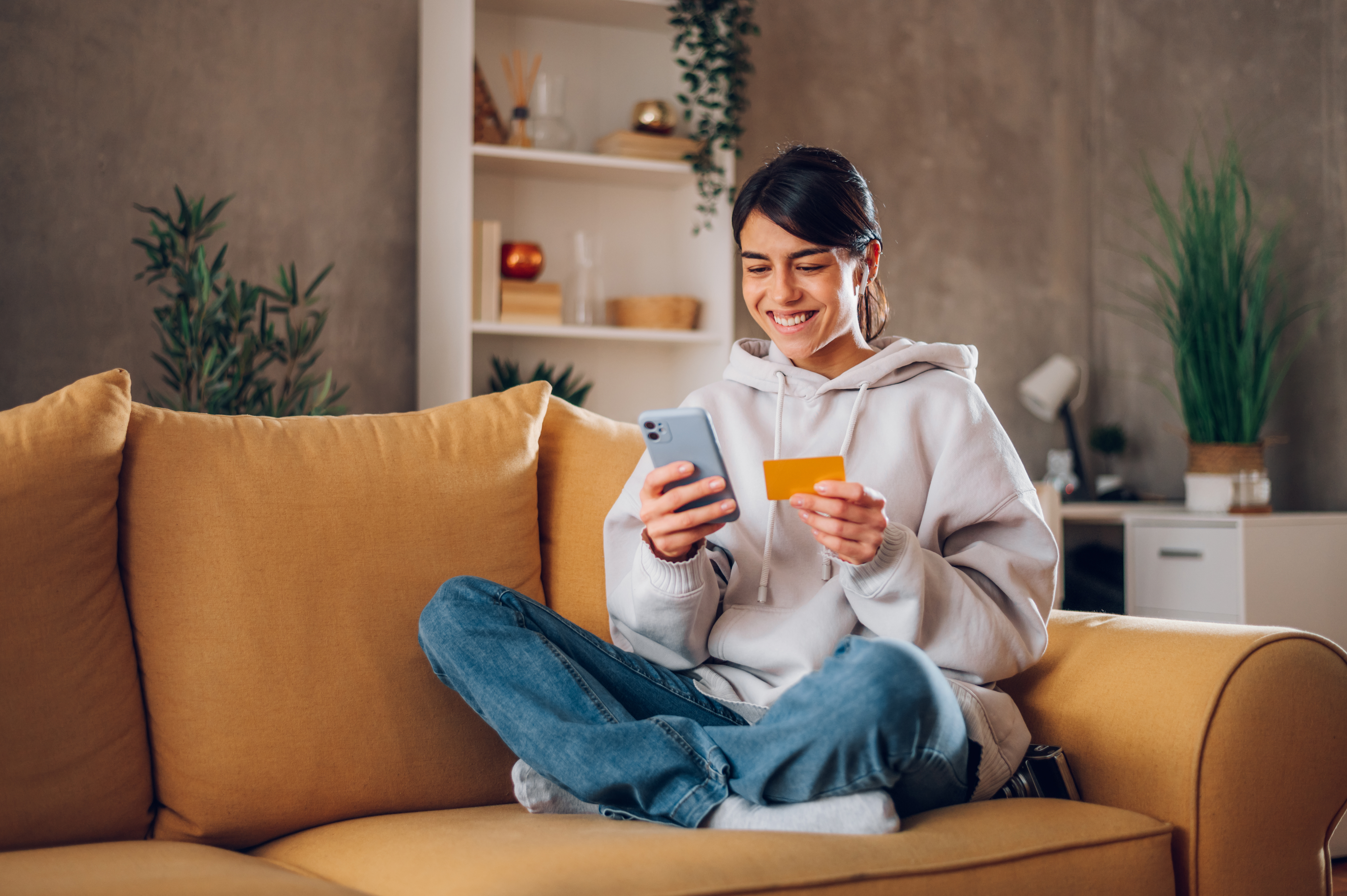 Woman sitting on couch, smiling, looking at phone and holding payment card