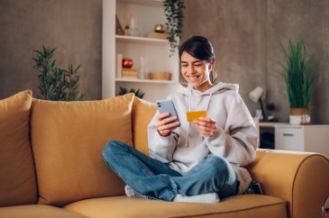 Woman sitting on couch, smiling, looking at phone and holding payment card