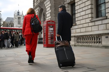 A woman with a backpack and a man with a suitcase are walking on a London street with a red telephone booth in the background.