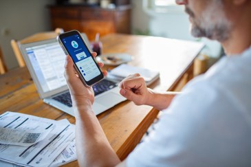 Man looking at cell phone, personal account, laptop on table