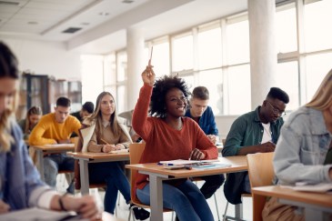 A young Black woman raises her hand in a college class; it's a sunlit room with modern desks that sit two students each.