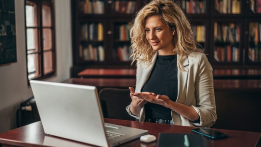 Woman sitting at desk wearing blazer and black shirt, engaging with someone on laptop; online meeting
