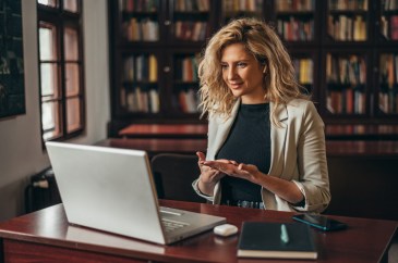 Woman sitting at desk wearing blazer and black shirt, engaging with someone on laptop; online meeting