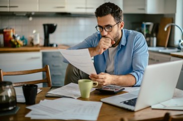Man sitting at kitchen table with hand on mouth, looking at paper, worried, calculator on table