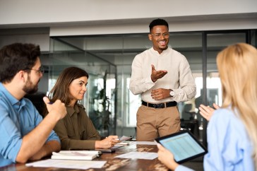 Group of employees sitting at table, man standing, talking and smiling