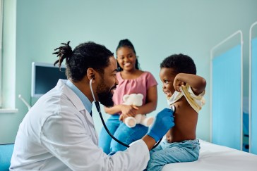Doctor listening to little boy's heart, mom in background, smiling