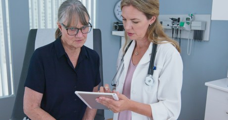 Older female patient sitting on exam table looking at tablet with female doctor