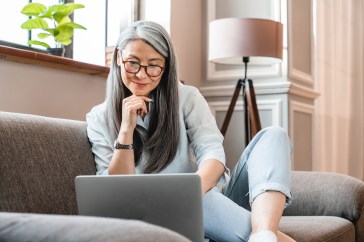 Woman sitting on couch, long gray hair and glasses, looking at computer
