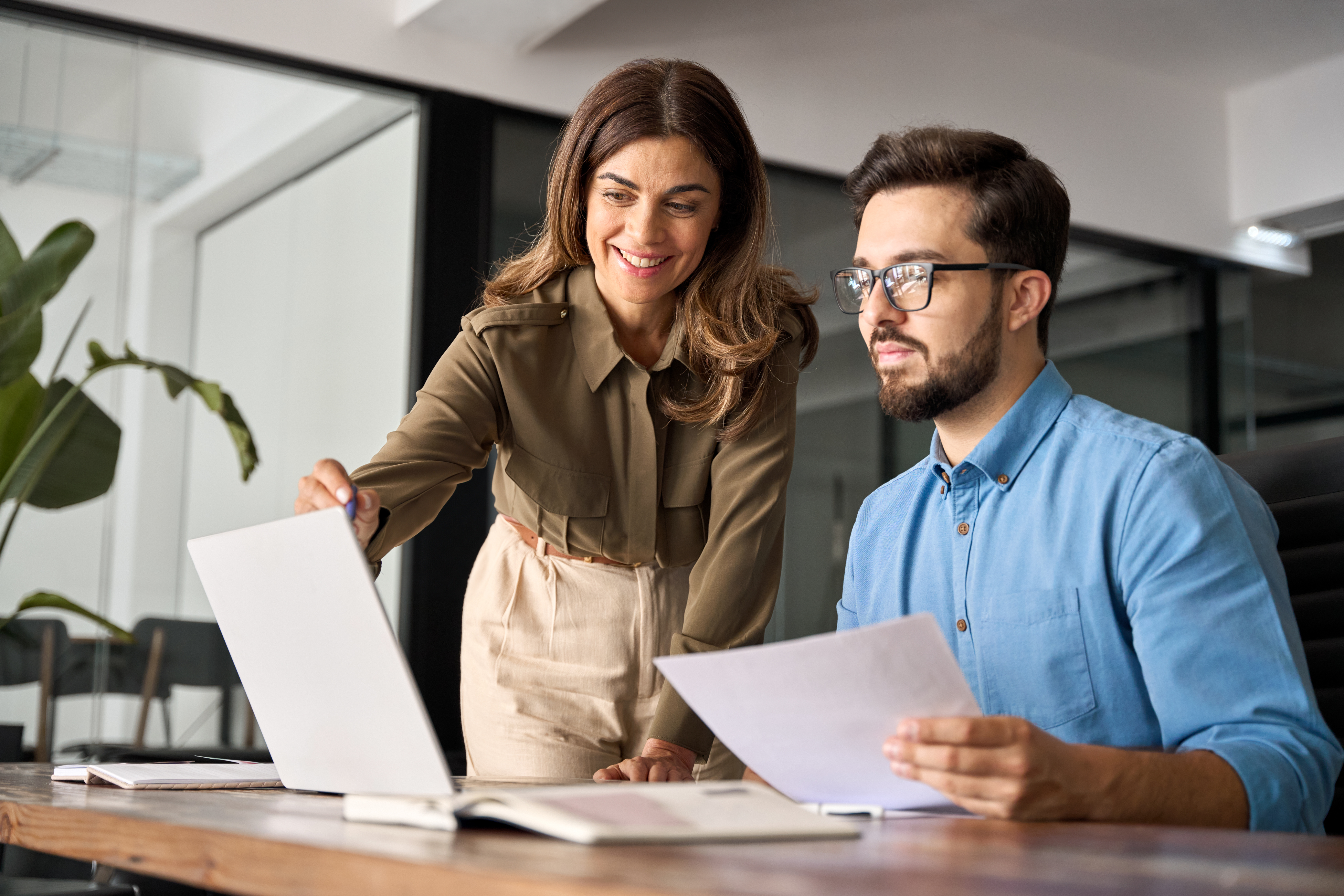 Male employee sitting at desk looking at computer and holding paper; female employee standing, pointing at computer, explaining