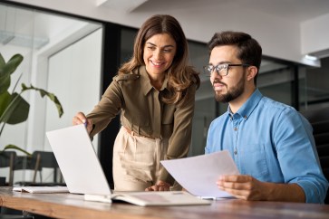 Male employee sitting at desk looking at computer and holding paper; female employee standing, pointing at computer, explaining