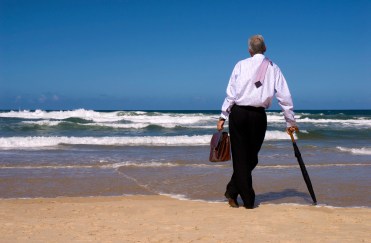 Man standing on beach, facing ocean, holding briefcase and umbrella with tie over shoulder