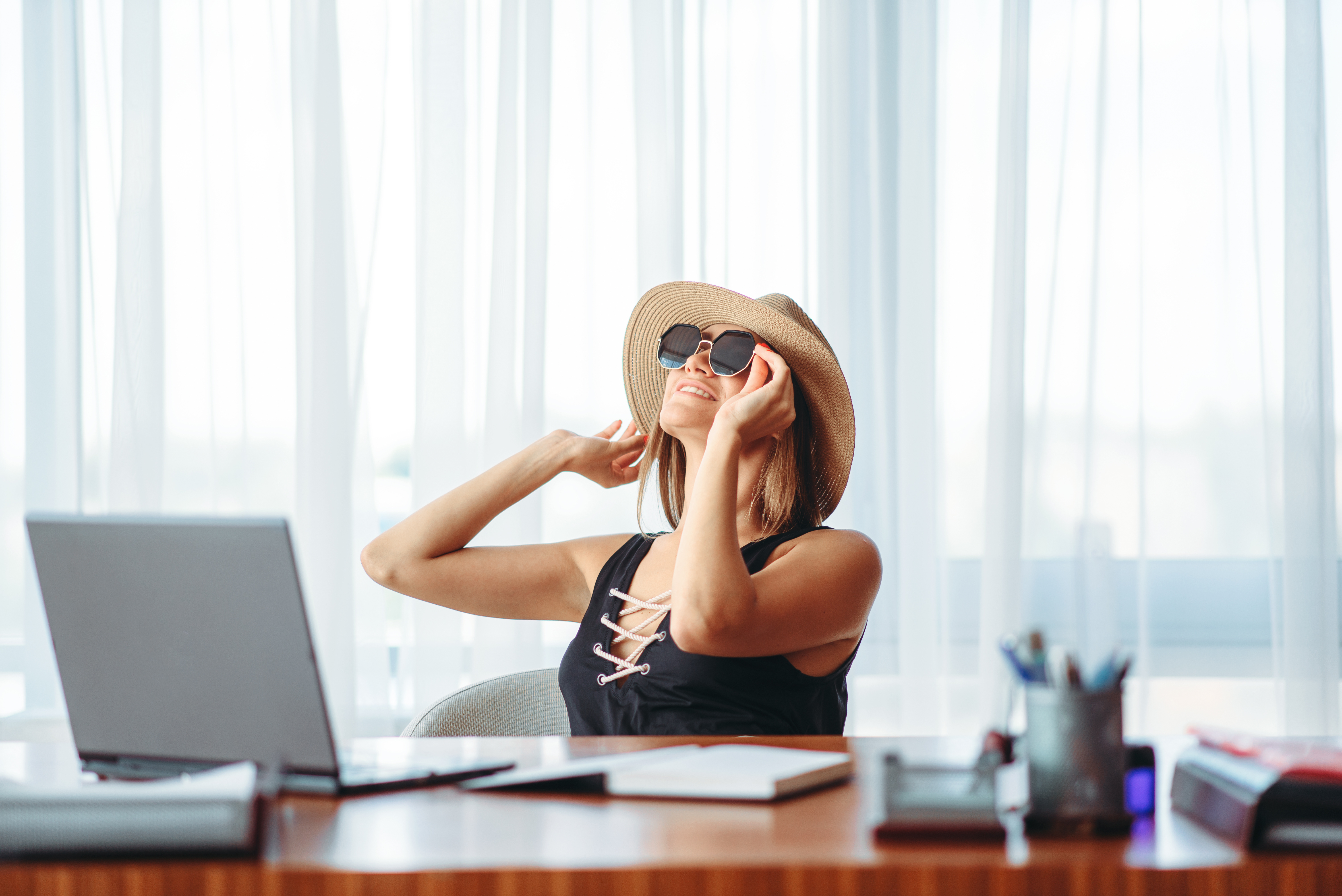 Woman sitting at desk thinking about vacation, wearing hat and sunglasses