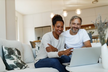 Couple sitting on couch looking at laptop, smilling, woman pointing at screen
