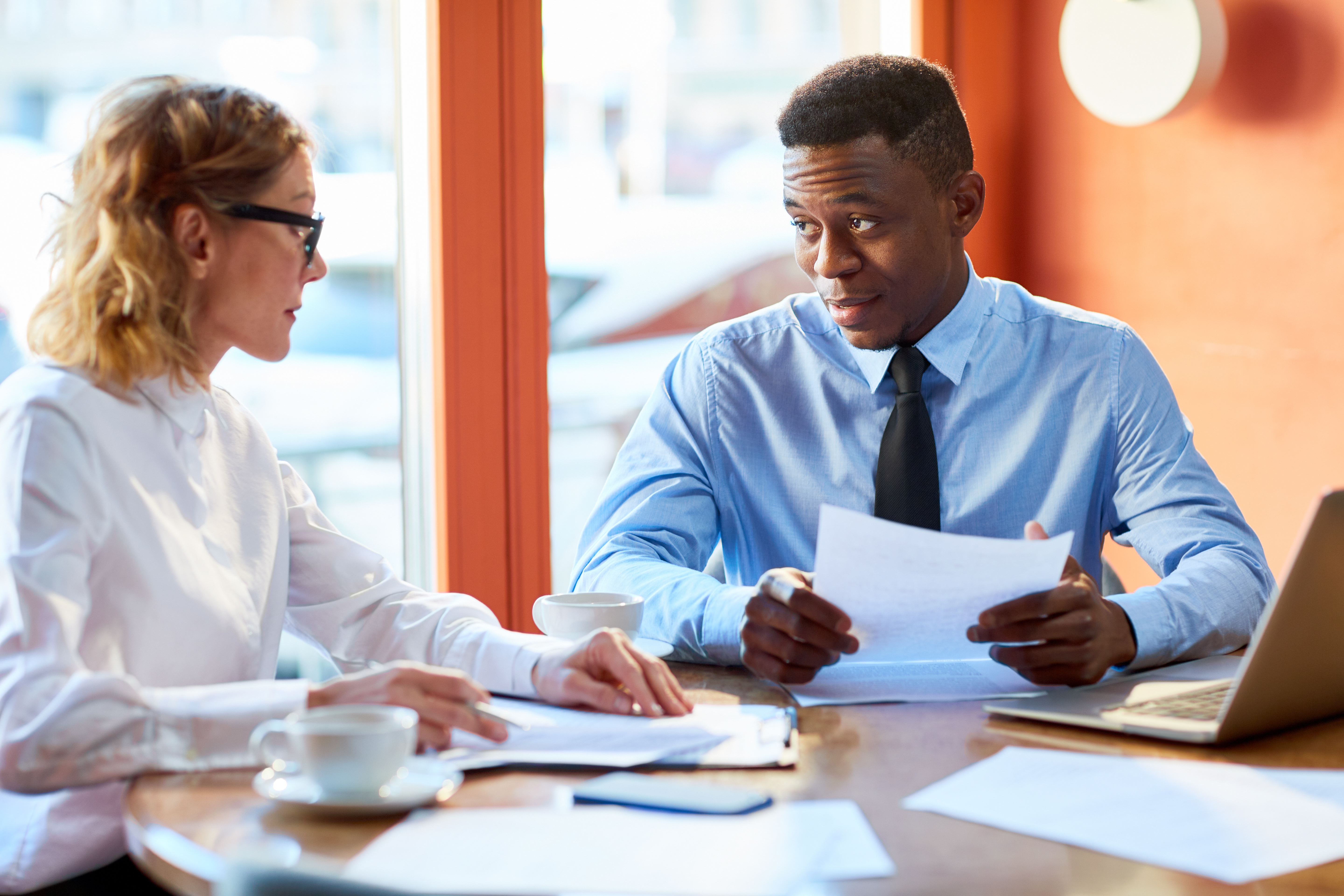 Two employees talking, sitting at table, looking at papers