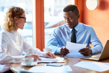 Two employees talking, sitting at table, looking at papers
