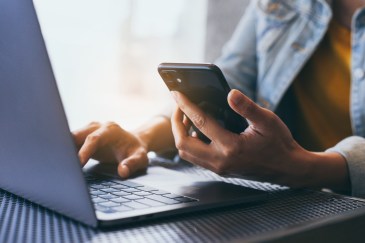 A person typing on a computer and holding a cellphone