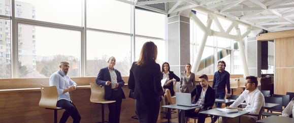 A woman speaks to a group of workers in an office building.