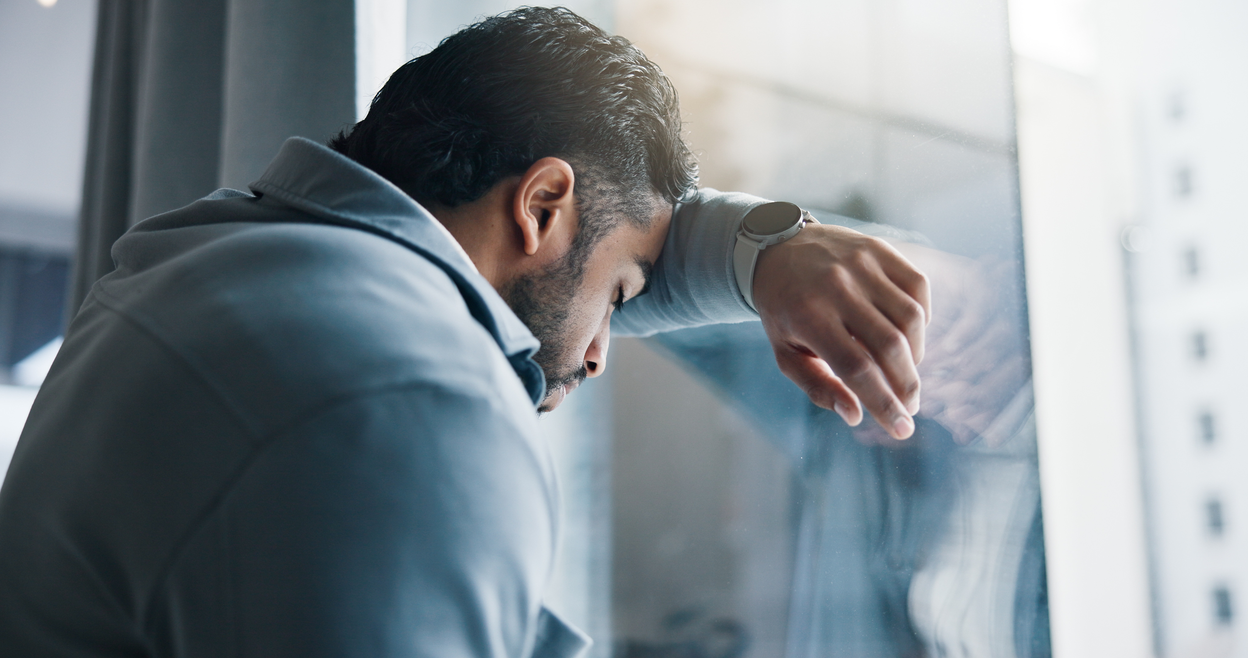 A man leans against a window with his head leaning against his forearm.