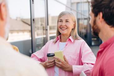 Woman with long gray hair, smiling, pink shirt, talking with two male coworkers