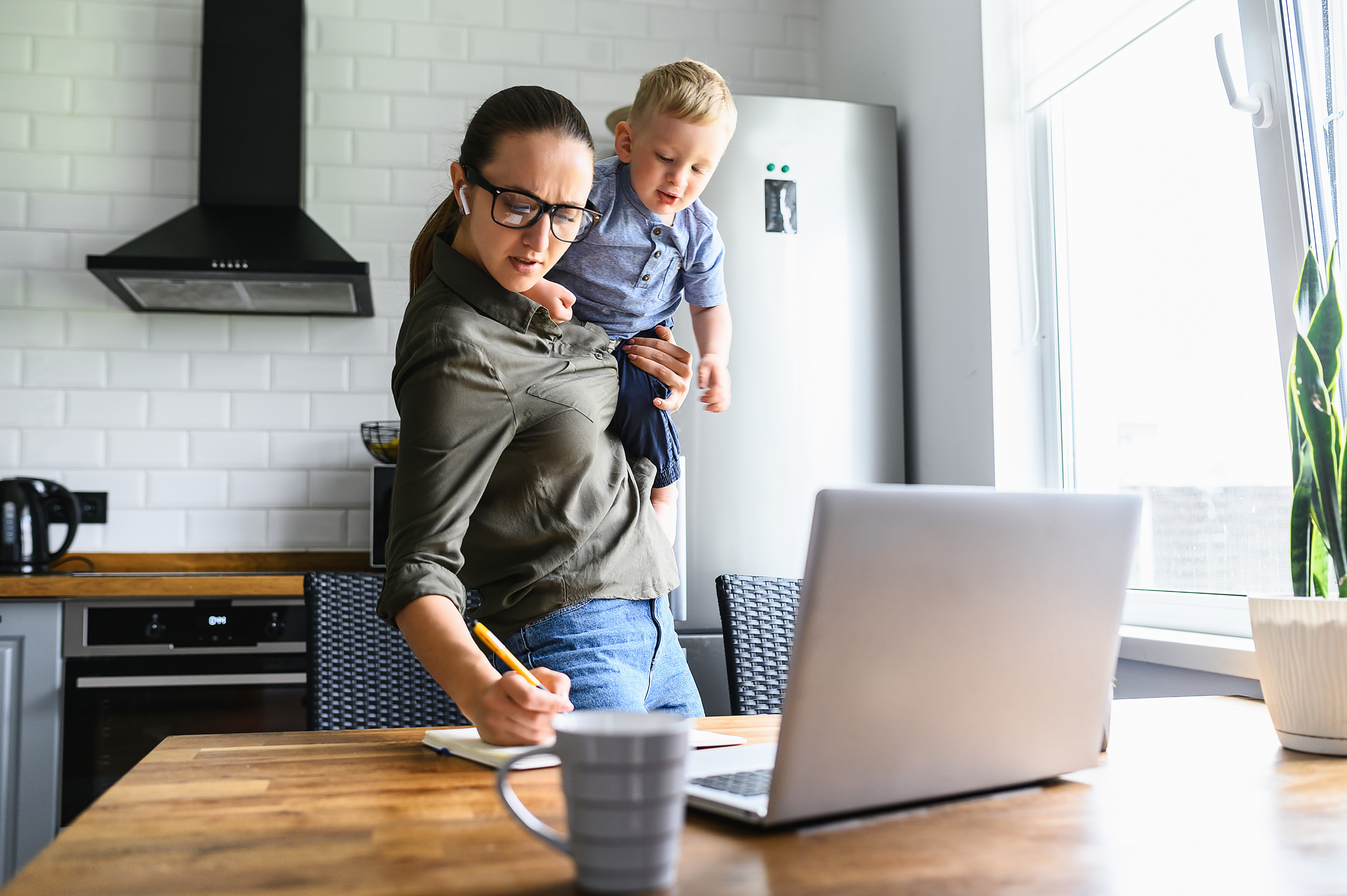 A mother holds her child while working at a laptop.