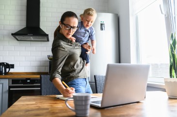 A mother holds her child while working at a laptop.