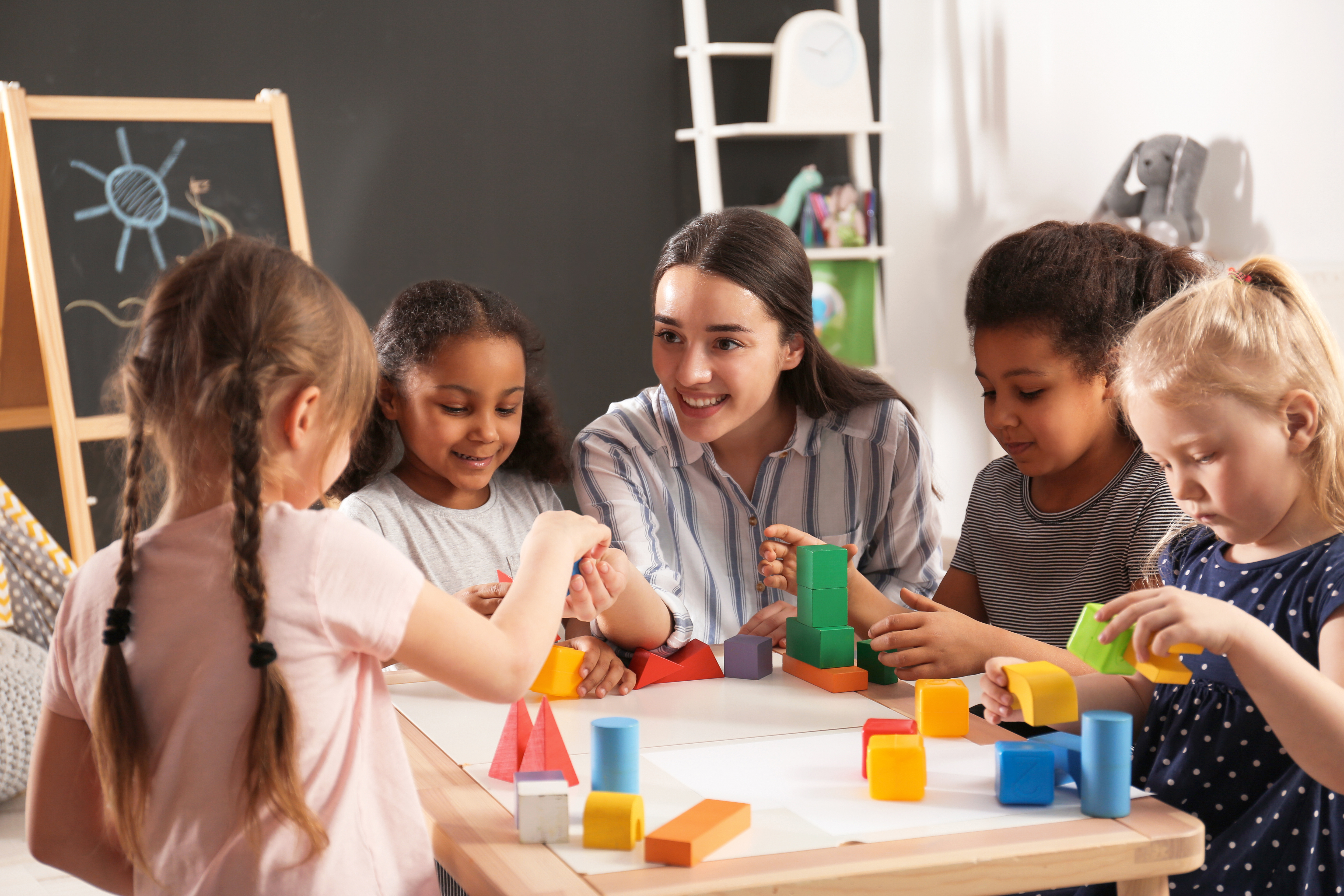 A group of kids play with blocks on a table while a caregiver interacts with them.