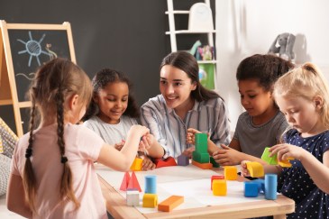 A group of kids play with blocks on a table while a caregiver interacts with them.