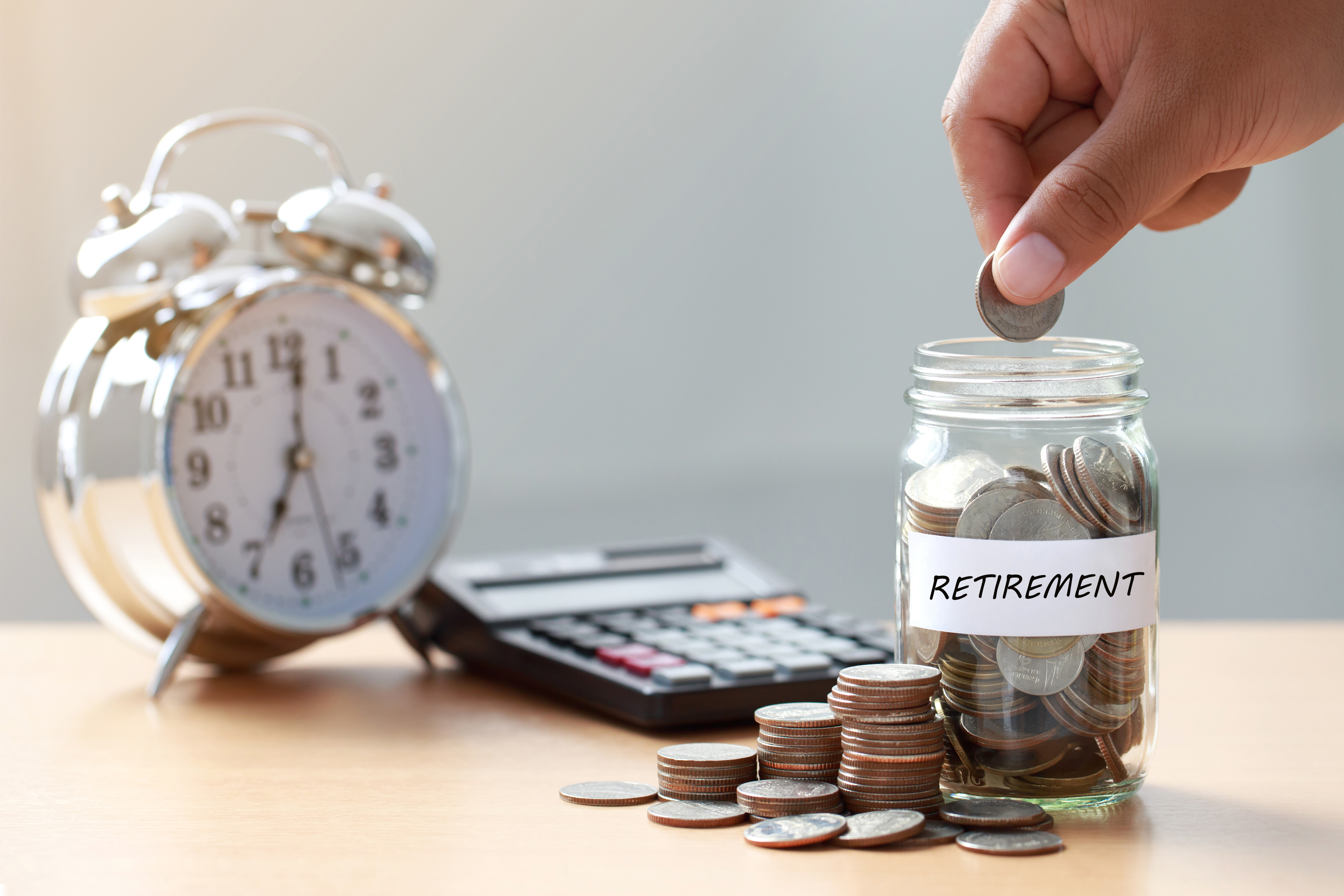 Hand putting money into jar labeled for retirement, next to clock and calculator