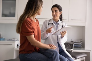 Professional Latina therapist holding modern digital tablet during conversation with woman patient, accessing electronic health record system, explain treatment plan or health test results.