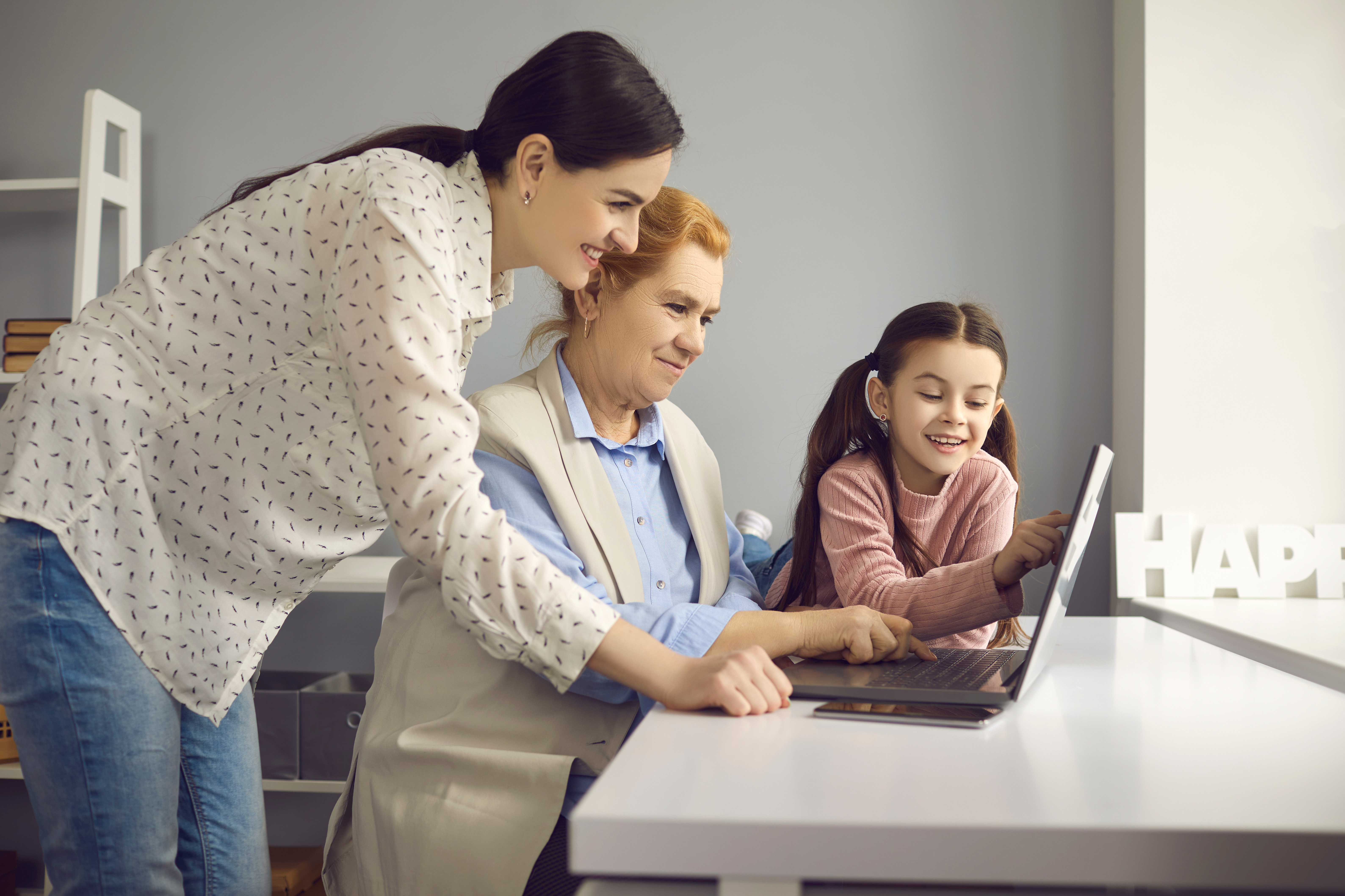 Older woman sitting at desk working on laptop, younger woman leaning over her shoulder, young girl watching