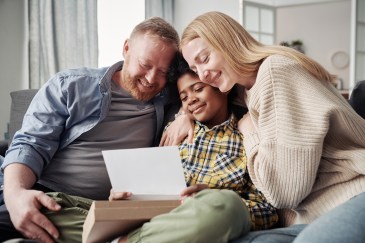 Happy parents embracing their African adoptive son while reading adoption agreement together on sofa.