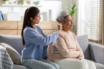 Young woman caring for elderly mother adjusting hearing aid device on couch at home, expressing warmth, love, compassion, and family connection in comfortable bright living room environment.