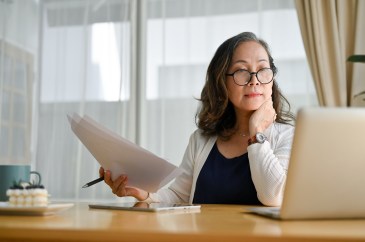 Woman holding papers looking at computer, wearing glasses