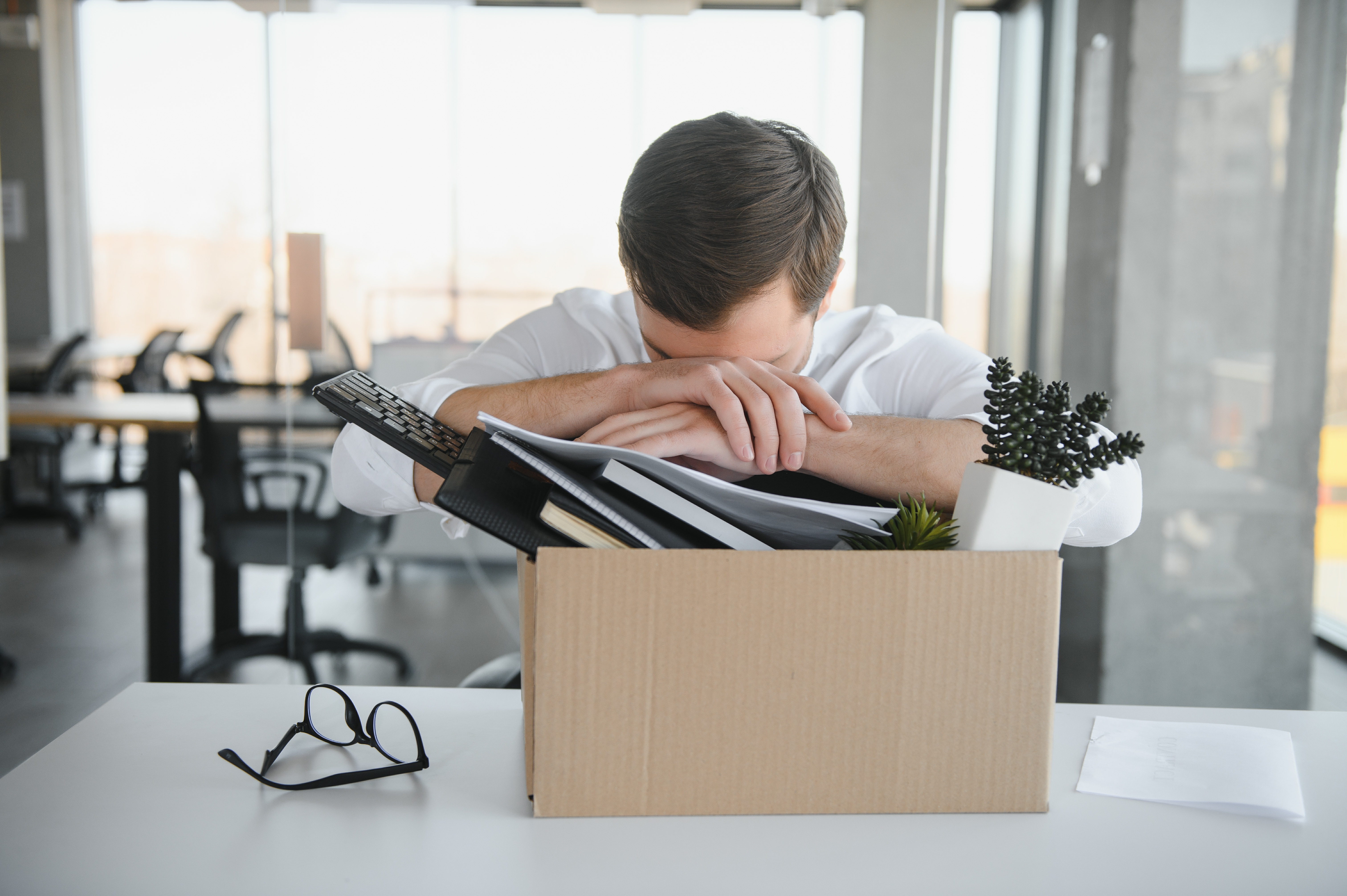 Man with head on arms, box packed with desk items, fired, laid off