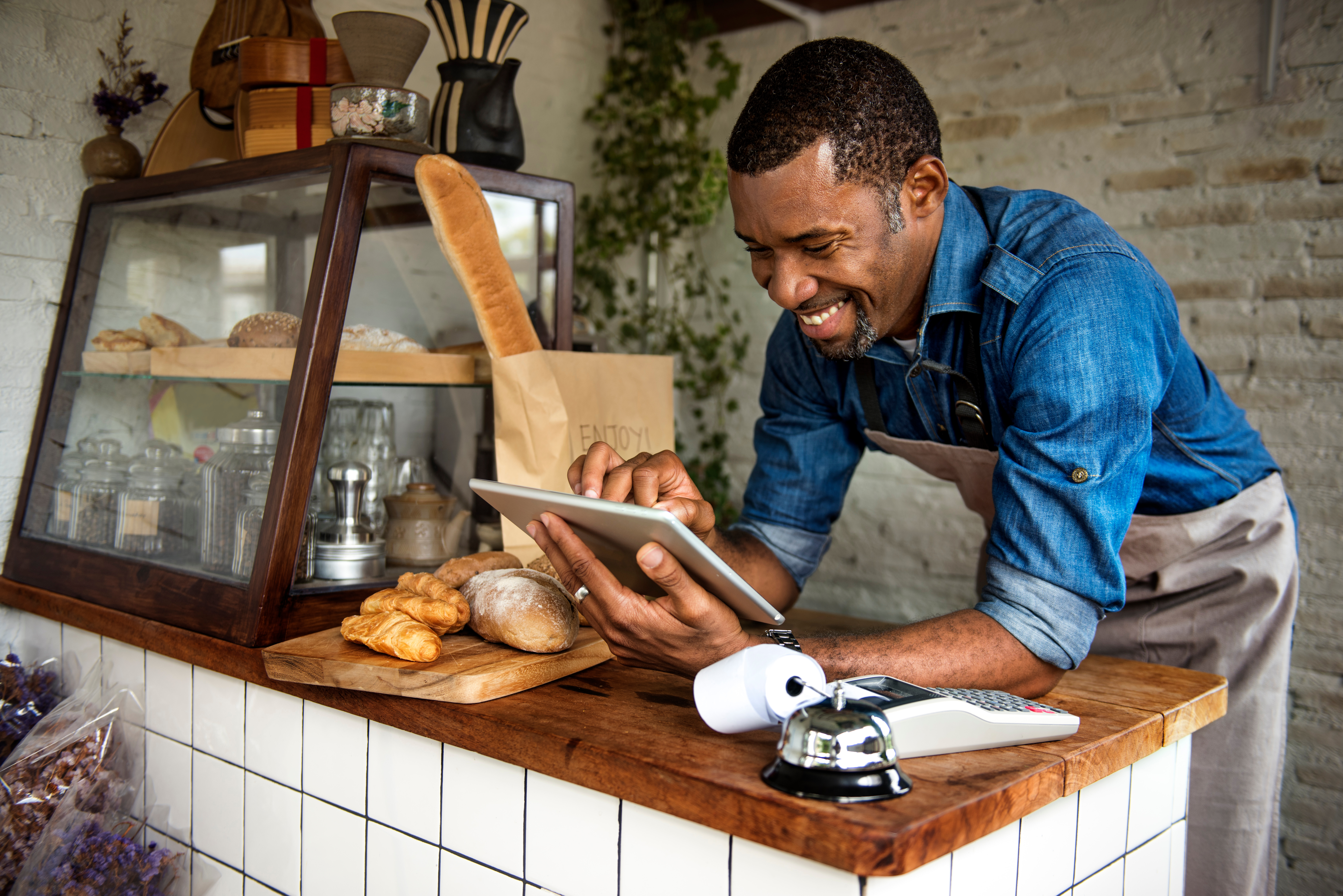 Man leaning on counter of bakery, employee, looking at tablet, smiling, small business