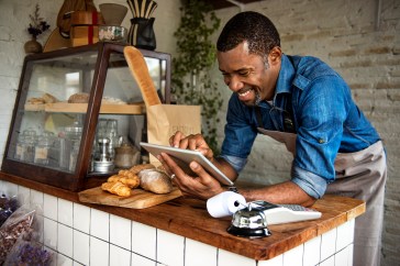 Man leaning on counter of bakery, employee, looking at tablet, smiling, small business