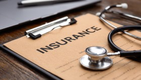 Medical stethoscope resting on brown insurance clipboard near laptop, highlighting healthcare expenses and medical documentation process.