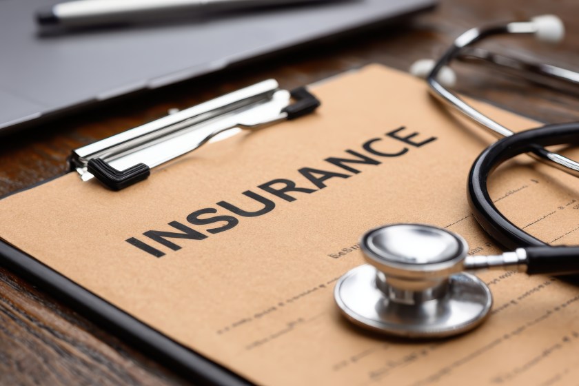 Medical stethoscope resting on brown insurance clipboard near laptop, highlighting healthcare expenses and medical documentation process.