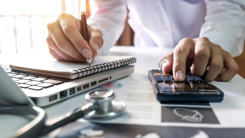 Healthcare costs and fees concept.Hand of smart doctor used a calculator and smartphone for medical costs at modern hospital in sun light.