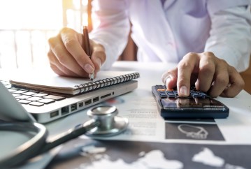 Healthcare costs and fees concept.Hand of smart doctor used a calculator and smartphone for medical costs at modern hospital in sun light.