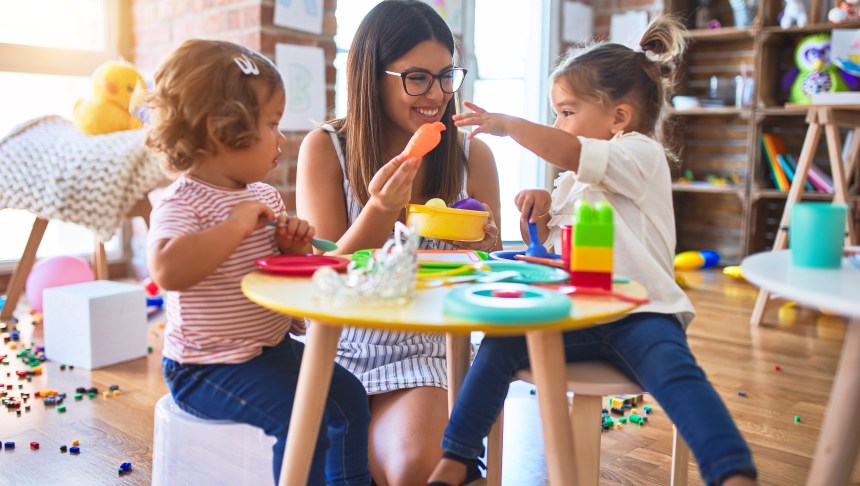 Young teacher and toddlers playing meals using plastic food and cutlery toy at kindergarten.