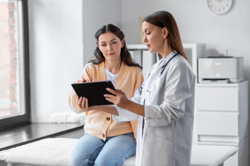 Woman sitting on table at doctor's office, speaking with doctor and looking at tablet