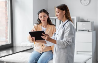 Woman sitting on table at doctor's office, speaking with doctor and looking at tablet