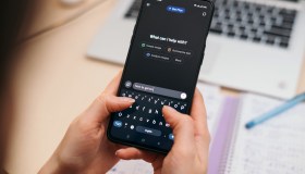 Person typing on smartphone using AI chat GPT app, with blurred notebook and laptop in background, modern workspace concept.