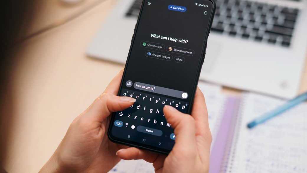 Person typing on smartphone using AI chat GPT app, with blurred notebook and laptop in background, modern workspace concept.